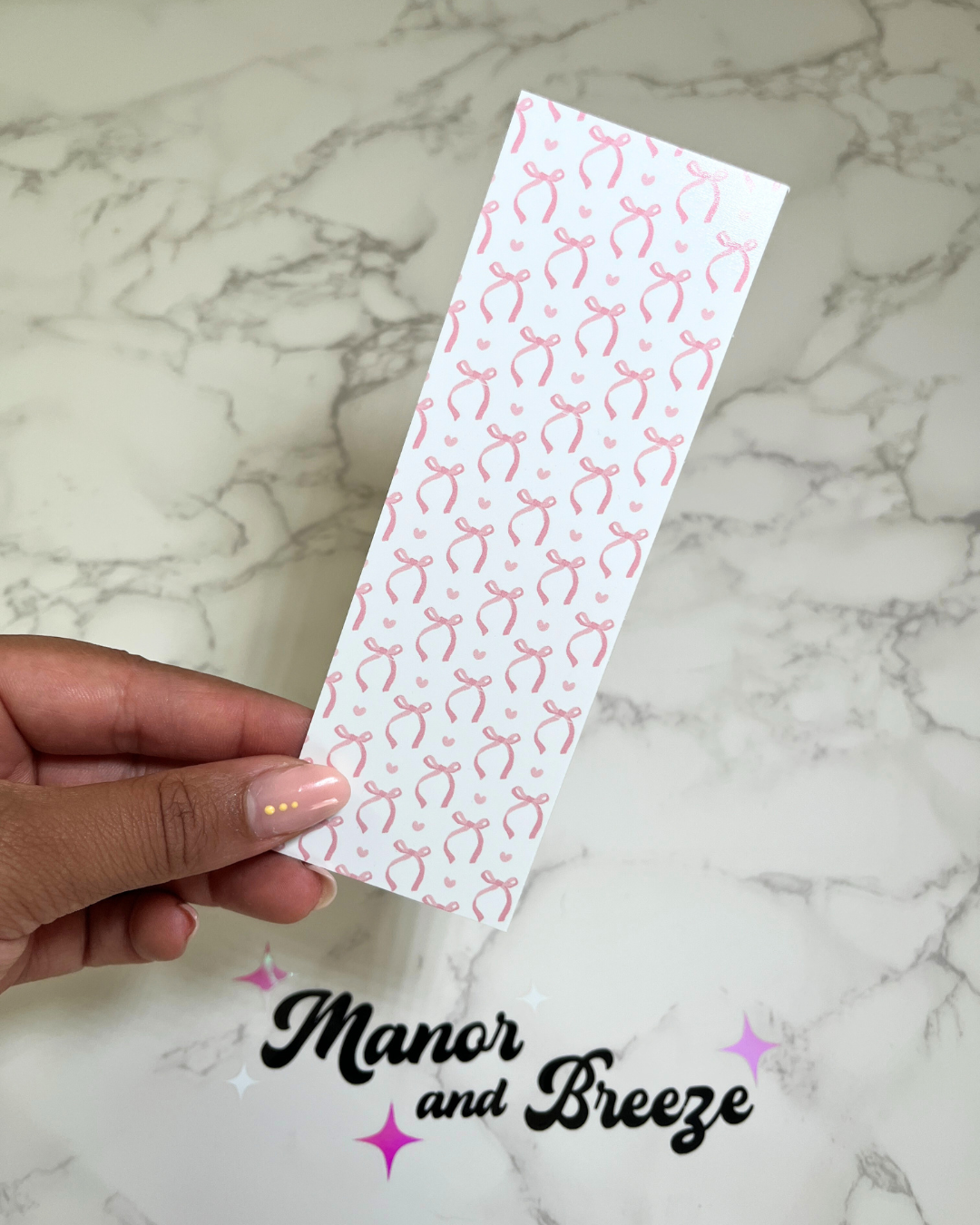 A hand holding a pink bookmark with a pattern of pink bows on a white background, placed on a marble surface.