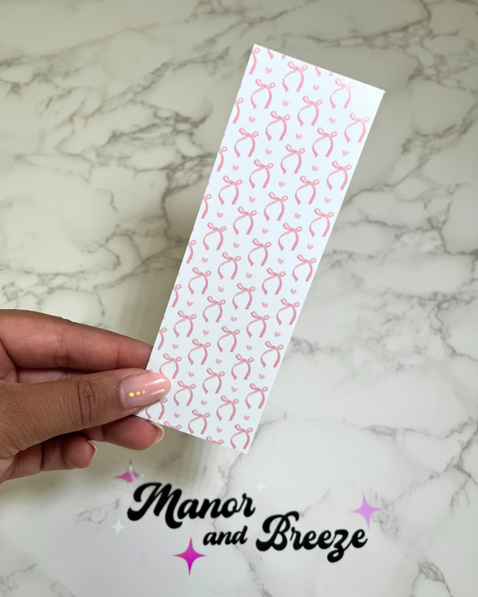 A hand holding a pink bookmark with a pattern of pink bows on a white background, placed on a marble surface.