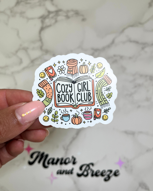 A sticker with a book design, including a graphic of a girl holding a book, and the text 'COZY GIRL BOOK CLUB'.
