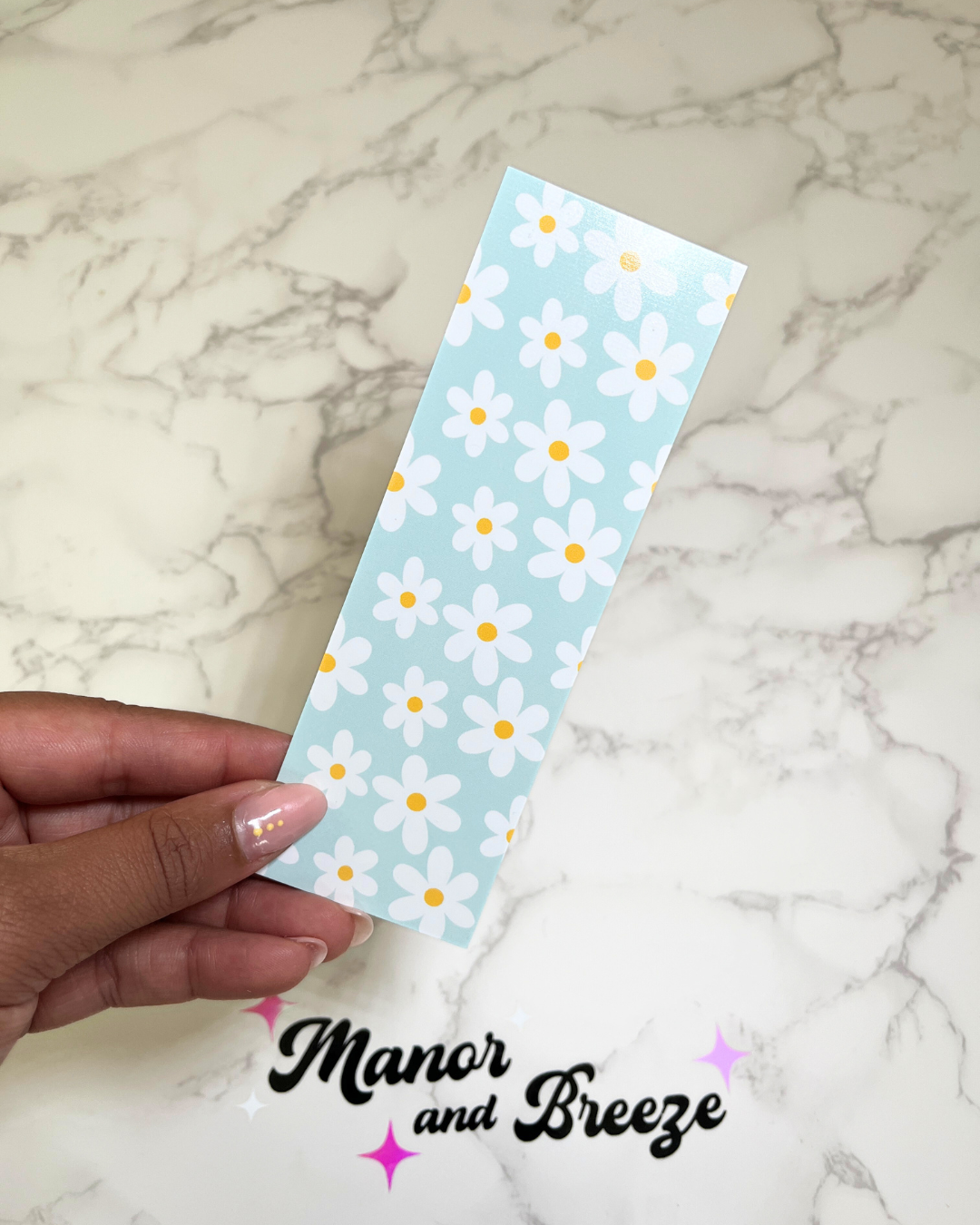 A bookmark with a blue background featuring a floral pattern of daisies, held between fingers against a marble background with 'Manor and Breeze' written below.