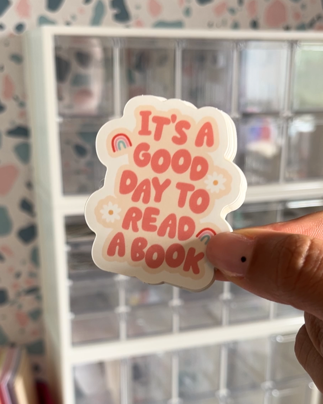 A hand holding a sticker with the text 'It's a good day to read a book' and book-related illustrations such as rainbows and flowers against a patterned background.