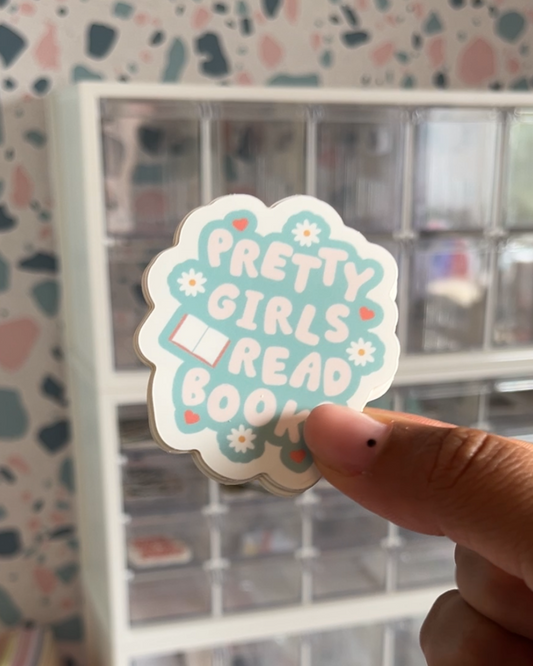 A sticker with the text 'Pretty Girls Read Books' held against a background that includes floral designs and a graphic of a book.